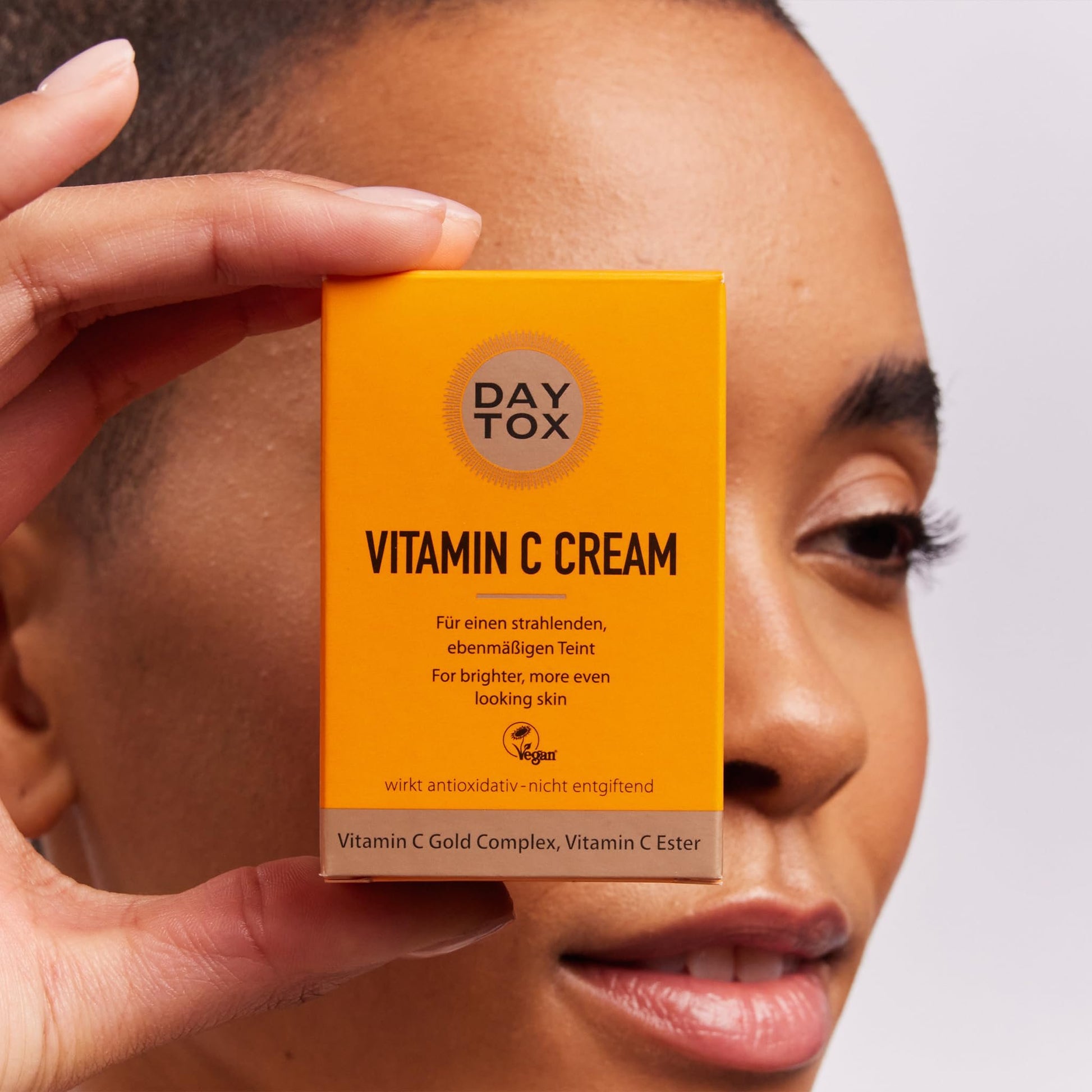 Eine Person hält die orangefarbene Produktverpackung der DAYTOX Vitamin C Creme vor ihr Gesicht.