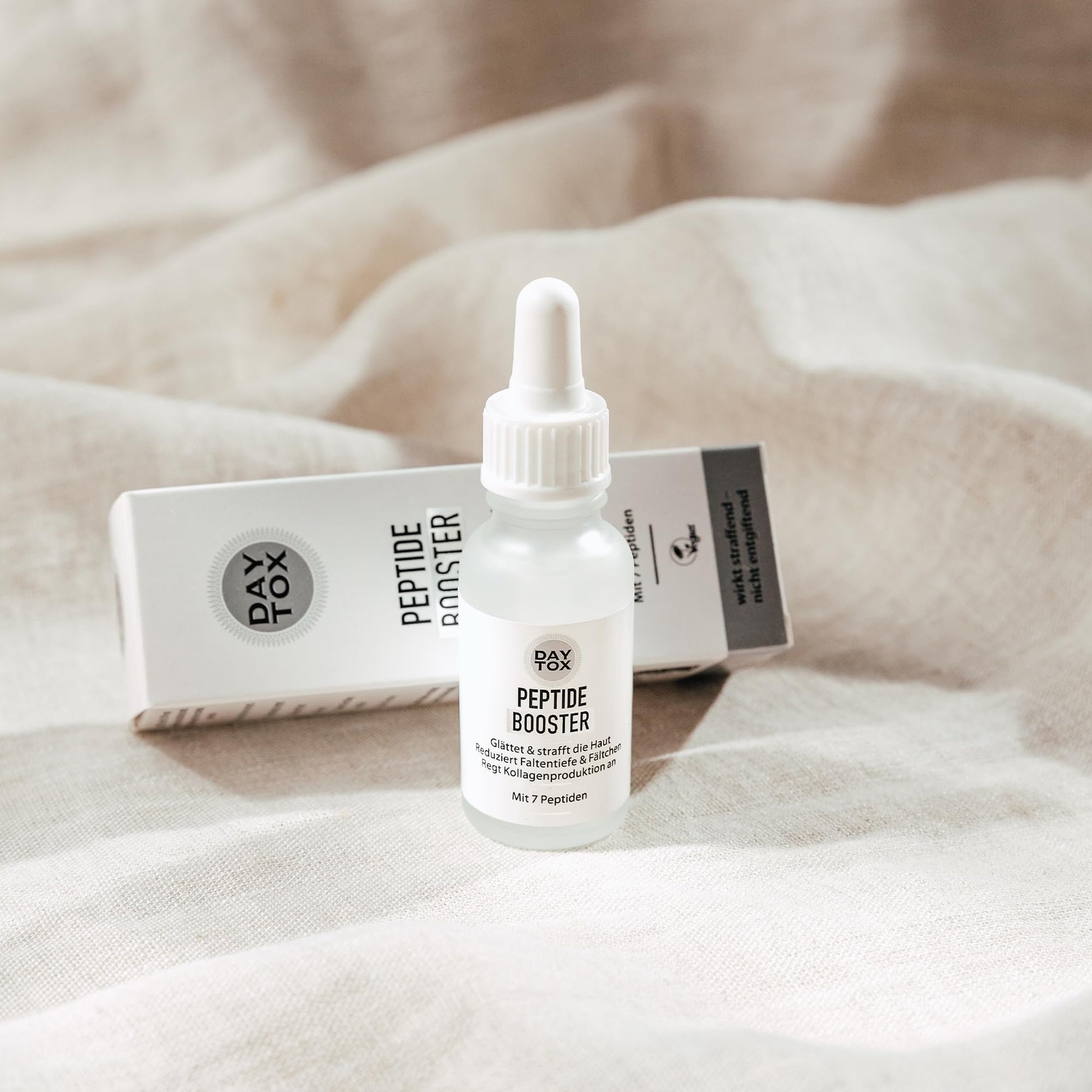 DAYTOX Peptide Booster Flasche mit Pipette liegt auf hellem Stoff. Die Produktverpackung steht dahinter.